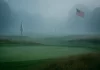 Campo de golf cubierto de niebla con bandera blanca en el green y la bandera estadounidense al fondo